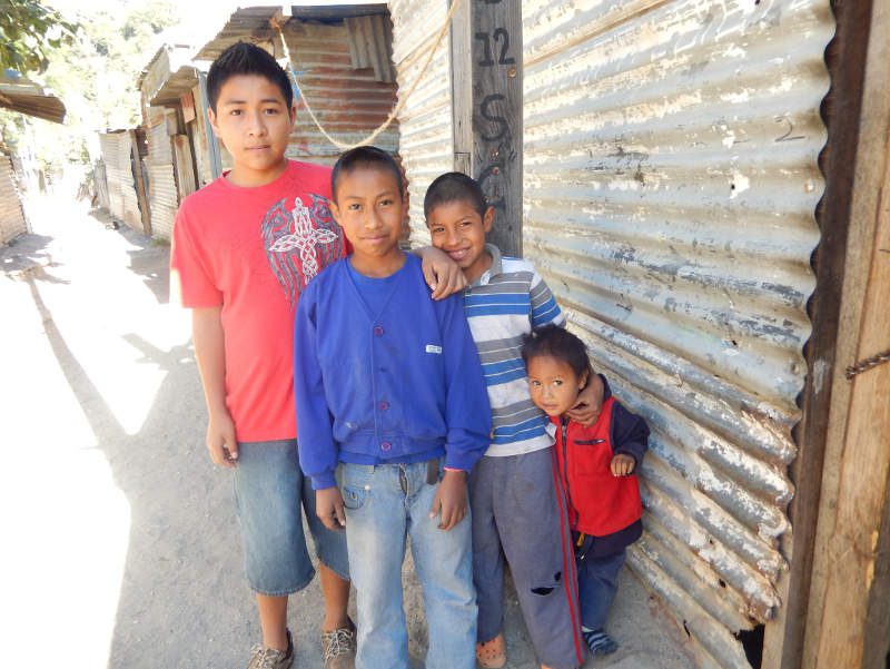 4 Guatemalan boys stand outside of a house made from old sheets of tin