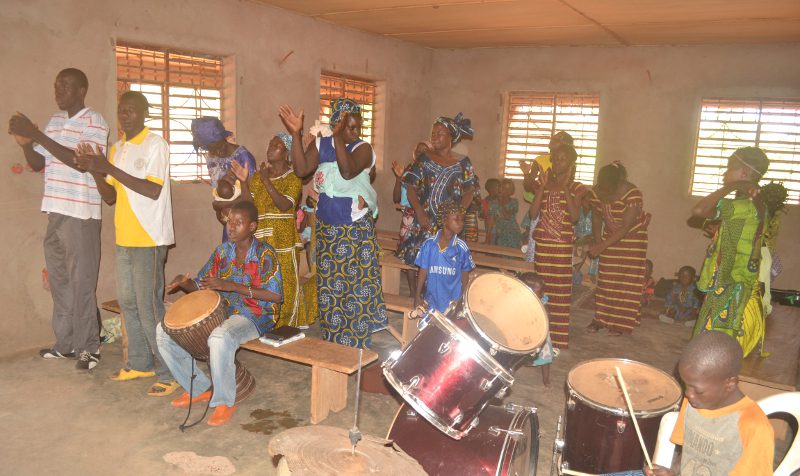 Burkinabé Christians stand in their church clapping their hands, one young man playing drums, and singing praises to God