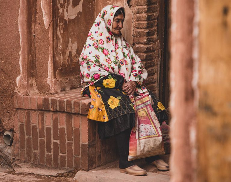 Elderly Iranian woman sits outside on a brick bench made built into a wall