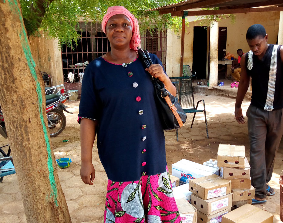 Malian woman stands holding her purse in front of a stack of boxes containing food and water