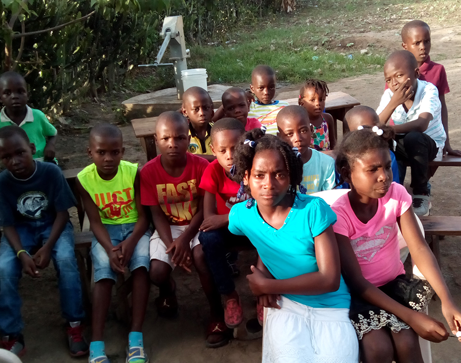 Haitian children sit on wooden picnic tables with a communal water pump behind them