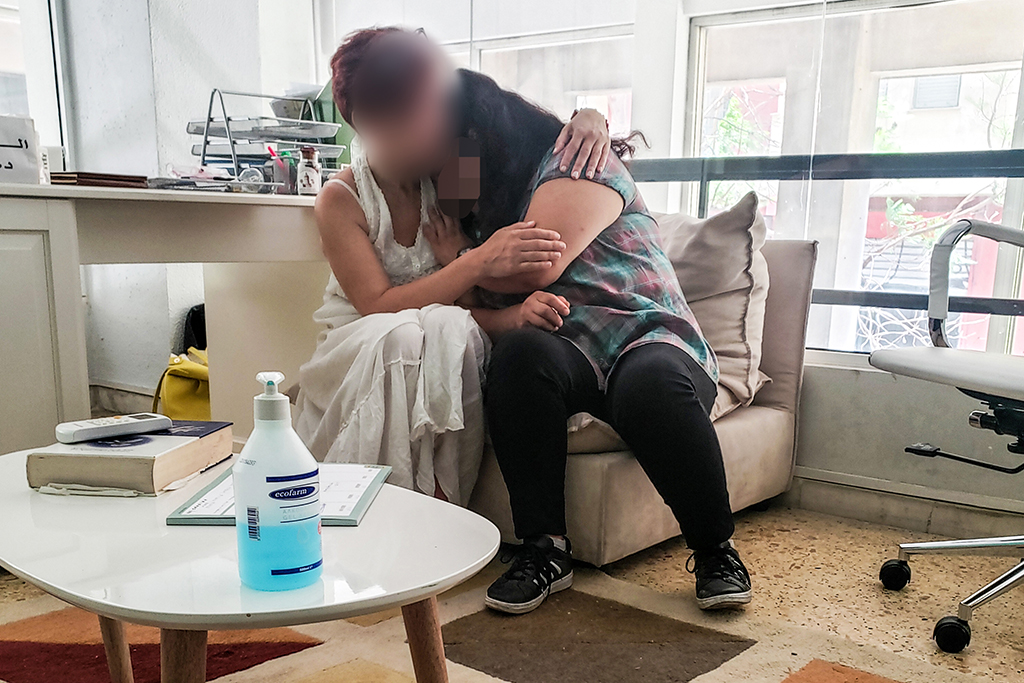 Greek women sits on a couch hugging a Middle Eastern refugee woman