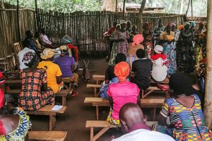 Burundian women stand singing while others sit on wooden benches listening