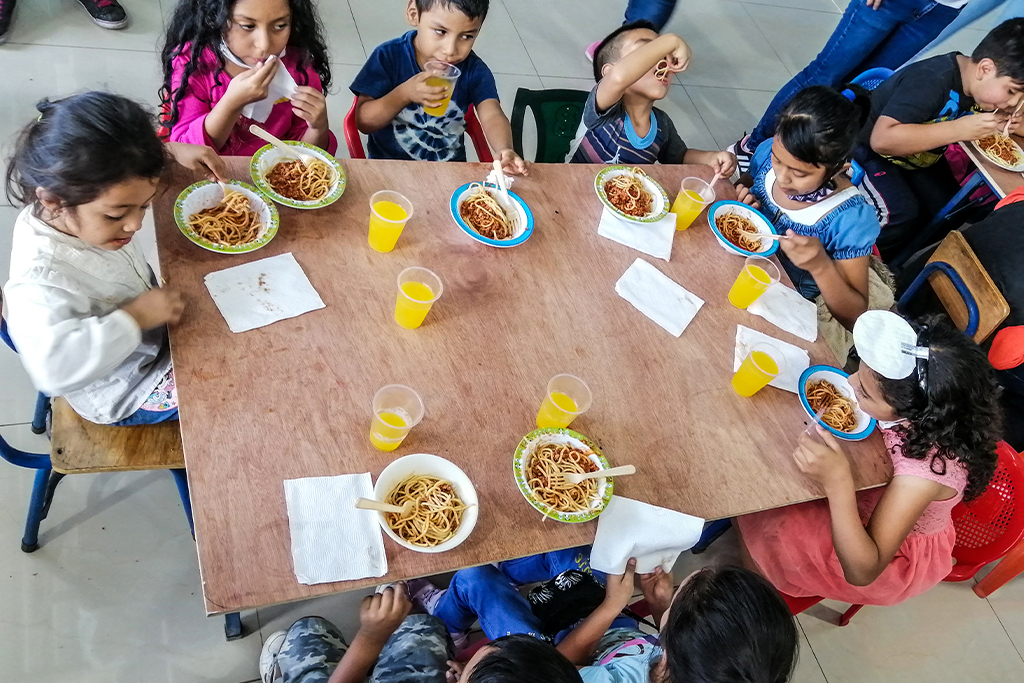 Guatemalan school children eating spaghetti and orange juice for lunch around a wooden table