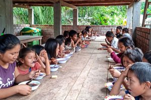 Peruvian children sit under a pavilion at a row of tables eating lunch