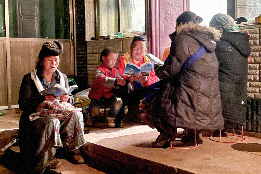 A group of Chinese Christian women wearing coats sit on stools while reading the Bible together