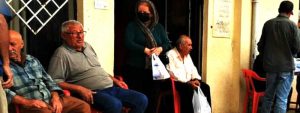 An Iranian woman walks out of a shop with a grocery bag while men sit outside in chairs on the street