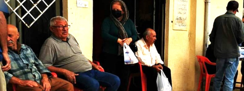 An Iranian woman walks out of a shop with a grocery bag while men sit outside in chairs on the street