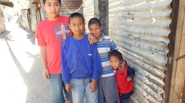 4 Guatemalan boys stand outside of a house made from old sheets of tin