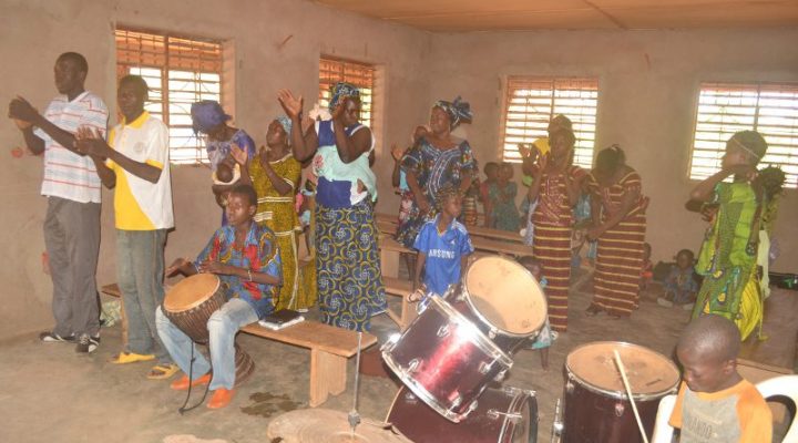 Burkinabé Christians stand in their church clapping their hands, one young man playing drums, and singing praises to God