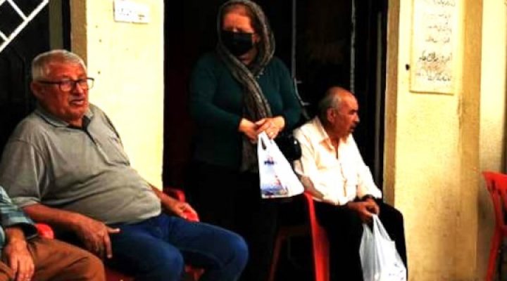 An Iranian woman walks out of a shop with a grocery bag while men sit outside in chairs on the street